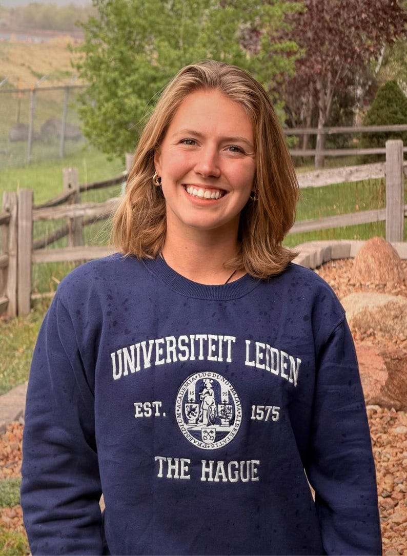Puede incluir: Una mujer con una sudadera azul marino con las palabras "UNIVERSITEIT LEIDEN" y "THE HAGUE" impresas en blanco. La sudadera tiene un emblema circular con texto y una figura. La mujer sonr&iacute;e, con un fondo natural al aire libre.
