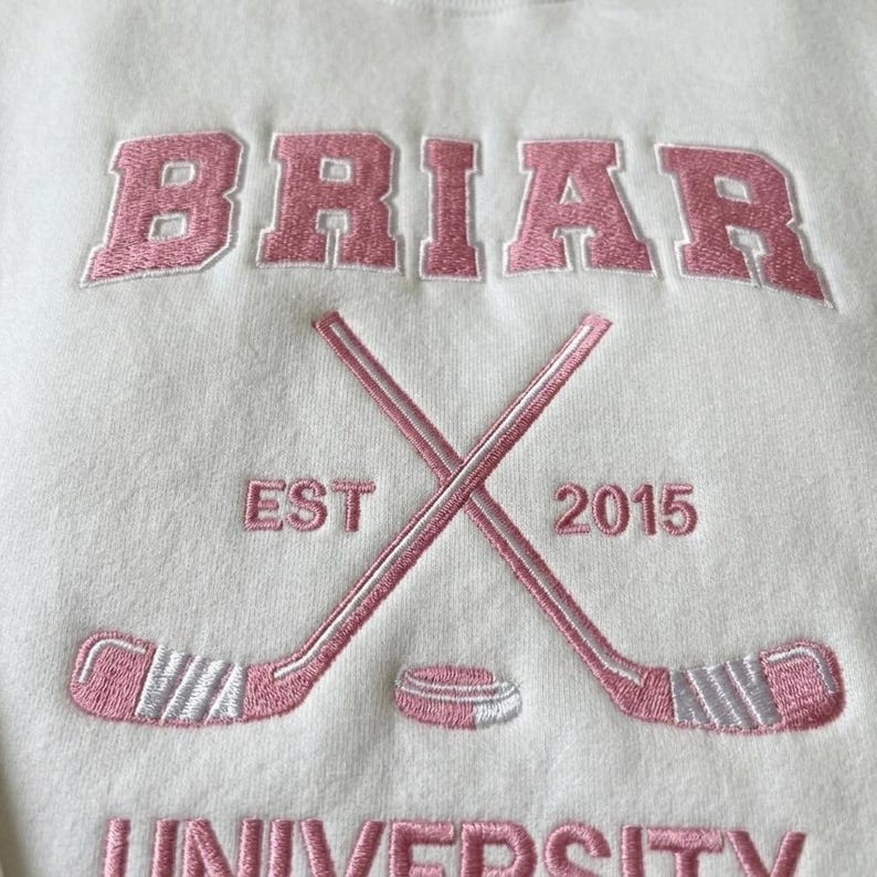 May include: Cream-colored sweatshirt with pink embroidered text and graphics. The word "BRIAR" is arched across the top. Below are crossed hockey sticks, a puck, and the text "EST 2015". The word "UNIVERSITY" is at the bottom.