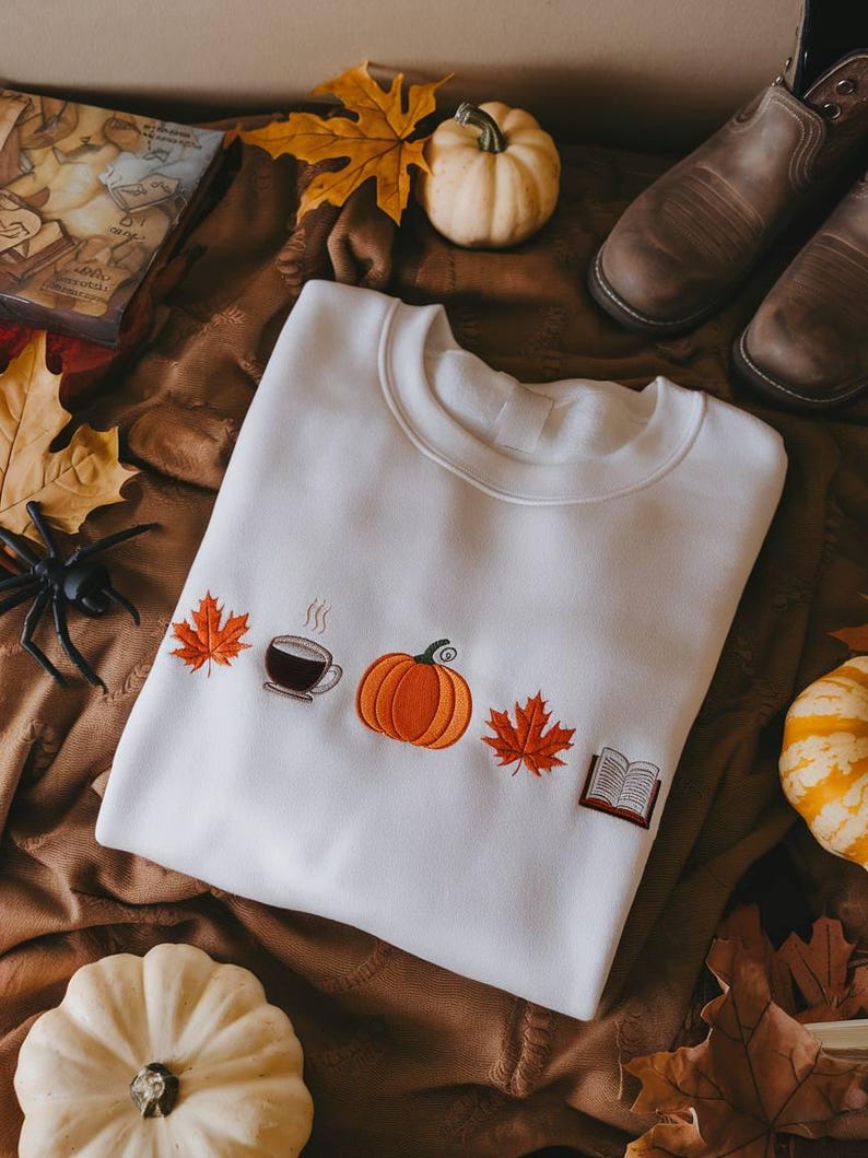 May include: Cream-coloured sweatshirt featuring embroidered autumnal motifs: a pumpkin, coffee cup, maple leaves, and a book. The image also shows pumpkins, leaves, and brown boots, creating a fall aesthetic.