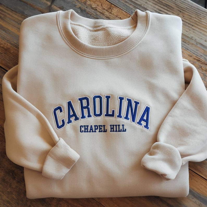 Op de afbeelding: Een cr&egrave;mekleurige sweatshirt met het woord "CAROLINA" in blauwe letters over de borst, met daaronder "CHAPEL HILL". De sweatshirt heeft een ronde hals en geribde manchetten.