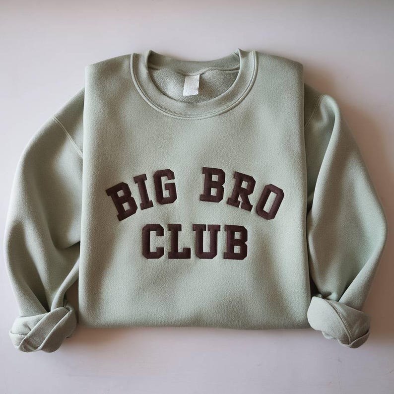 Puede incluir: Una sudadera verde salvia con las palabras "BIG BRO CLUB" en letras oscuras de bloque marr&oacute;n. La sudadera est&aacute; doblada, con las mangas dobladas. La sudadera de cuello redondo est&aacute; hecha de un material suave.