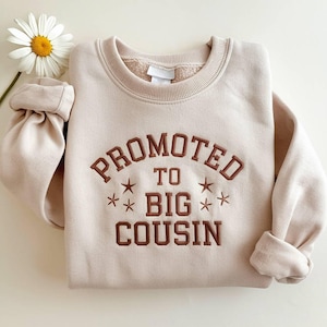 Può includere: Una felpa beige con la scritta ricamata "PROMOTED TO BIG COUSIN" in marrone. Il disegno include stelle. Un fiore di margherita è nell'angolo in alto a sinistra. Le maniche sono piegate.