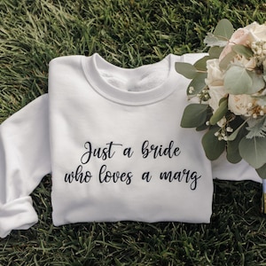 May include: A white sweatshirt with the text "Just a bride who loves a marg" in black script. A bouquet of white and pink flowers with greenery rests on the right side of the sweatshirt, all on a bed of green grass.