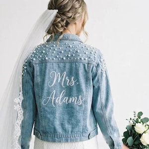 Puede incluir: Chaqueta vaquera azul claro adornada con perlas de imitación en los hombros. La parte trasera de la chaqueta presenta el texto bordado "Mrs. Adams" en escritura blanca. Un velo de novia es visible en el lado izquierdo de la imagen.