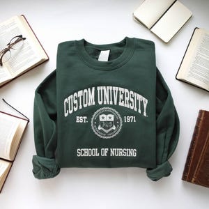 Könnte beinhalten: Ein dunkelgrüner Pullover mit dem Text "Custom University" in Weiß, einem Wappen mit dem Text "Est. 1971" und dem Text "School of Nursing" in Weiß.