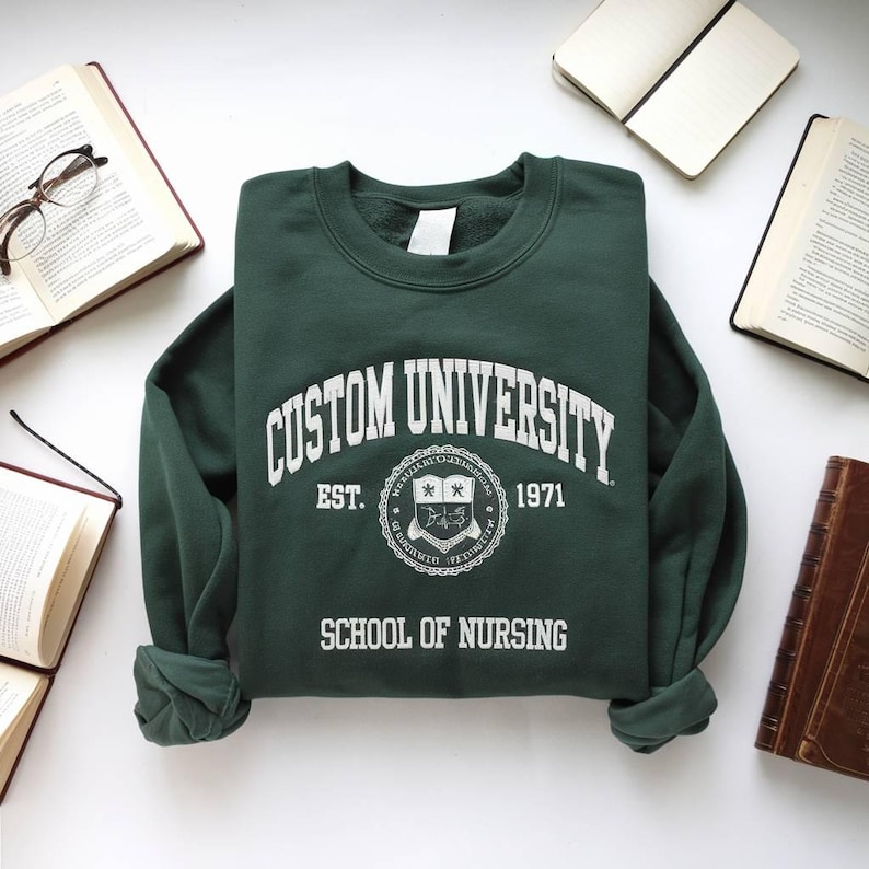 Puede incluir: Una sudadera verde oscuro con la inscripci&oacute;n blanca "CUSTOM UNIVERSITY". Debajo, un escudo con las palabras "EST. 1971" y "SCHOOL OF NURSING". Libros abiertos y gafas est&aacute;n colocados alrededor de la sudadera.