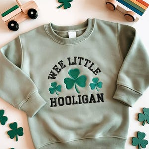 Könnte beinhalten: Salbeigrünes Sweatshirt mit dem Text "WEE LITTLE HOOLIGAN" und drei grünen Kleeblatt-Designs. Das Sweatshirt hat einen Rundhalsausschnitt und lange Ärmel. Holzspielzeugautos und Kleeblatt-Ausschnitte sind verstreut.