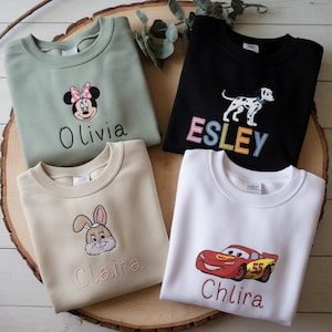 Może przedstawiać: Cztery spersonalizowane bluzy z haftowanymi wzorami. Bluzy są w kolorze zielonym, czarnym, beżowym i białym. Wzory obejmują Myszki Miki, dalmatyńczyka, królika i czerwony samochód wyścigowy. Na bluzach są wyhaftowane imiona Olivia, Esley i Chlira.