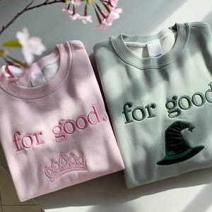 Puede incluir: Se muestran dos sudaderas, una rosa claro y otra verde salvia. La sudadera rosa tiene el texto bordado "for good." y una corona. La sudadera verde tiene el texto bordado "for good." y un sombrero de bruja.