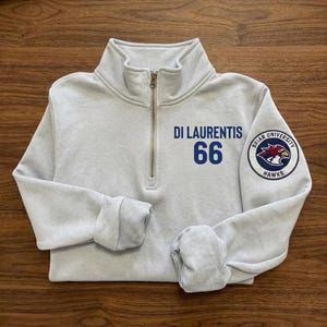 Könnte beinhalten: Hellgraues Sweatshirt mit Viertelreißverschluss und dem blauen Text "DI LAURENTIS 66". Ein Aufnäher auf dem Ärmel zeigt "Briar University Hawks" mit einem roten Falken-Emblem. Das Sweatshirt hat einen gerippten Kragen und Bündchen.