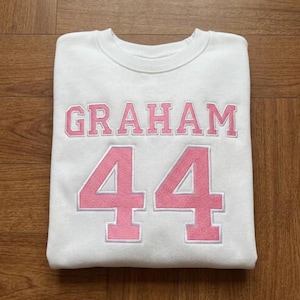May include: White sweatshirt with the name "GRAHAM" in pink block letters above the number "44" in pink. The sweatshirt is folded on a wooden surface.