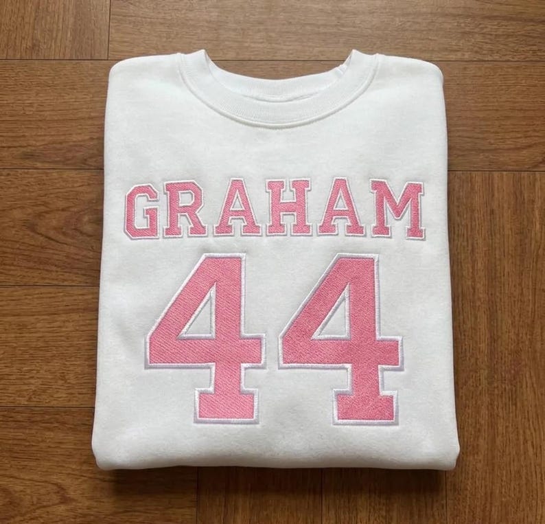 May include: White sweatshirt with the name "GRAHAM" in pink block letters above the number "44" in pink. The sweatshirt is folded on a wooden surface.