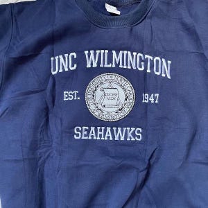 Puede incluir: Una sudadera azul marino con texto bordado en blanco que dice "UNC WILMINGTON" y "SEAHAWKS". Un emblema circular presenta texto y el a&ntilde;o "1947". La sudadera tiene cuello redondo y un corte holgado.