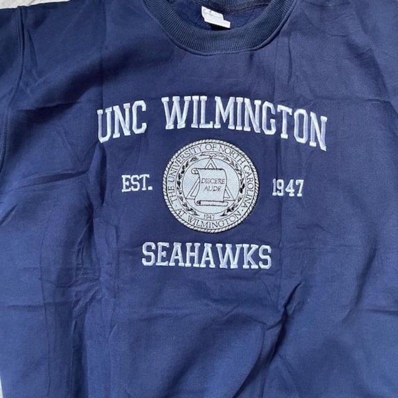 Puede incluir: Una sudadera azul marino con texto bordado en blanco que dice "UNC WILMINGTON" y "SEAHAWKS". Un emblema circular presenta texto y el a&ntilde;o "1947". La sudadera tiene cuello redondo y un corte holgado.