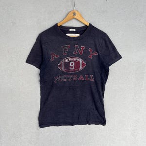 May include: A dark charcoal grey short-sleeve t-shirt featuring a distressed graphic print. The design includes the text AFNY arched above a football illustration with the number 9 inside, and the word FOOTBALL printed below in red and white.
