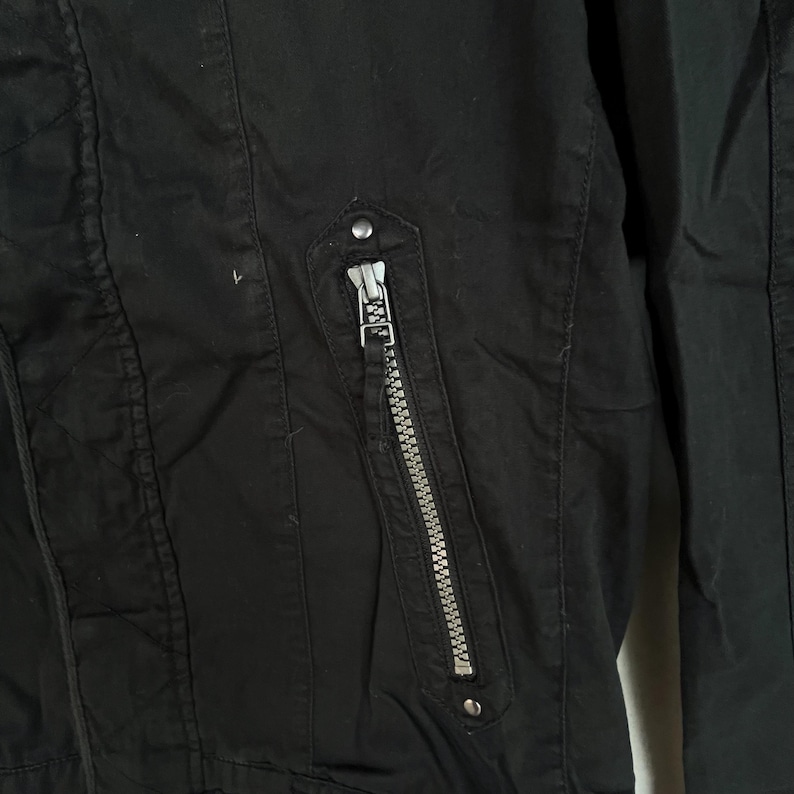 Puede incluir: Primer plano de una chaqueta negra con cremallera plateada y un peque&ntilde;o bot&oacute;n redondo plateado. La chaqueta tiene un dise&ntilde;o acolchado en el lado izquierdo. La cremallera est&aacute; centrada y es vertical.