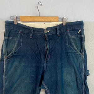 May include: Close-up of blue denim jeans with white stitching and red accents. The jeans are hanging on a wooden hanger. The jeans have a button closure and a zipper fly. The pockets have a unique design.