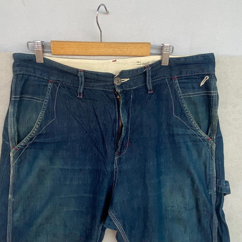 May include: Close-up of blue denim jeans with white stitching and red accents. The jeans are hanging on a wooden hanger. The jeans have a button closure and a zipper fly. The pockets have a unique design.