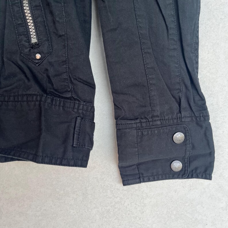 Puede incluir: Primer plano de una chaqueta negra con cremallera plateada y dos botones plateados en el pu&ntilde;o. La chaqueta tiene un corte entallado con detalles de costura visibles y un peque&ntilde;o bolsillo.
