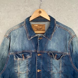 Pu&ograve; includere: Giacca di jeans blu dal design classico, con chiusura a bottoni, due tasche sul petto e colletto. La giacca &egrave; appesa a una gruccia di legno. Il denim ha un aspetto sbiadito e usurato.