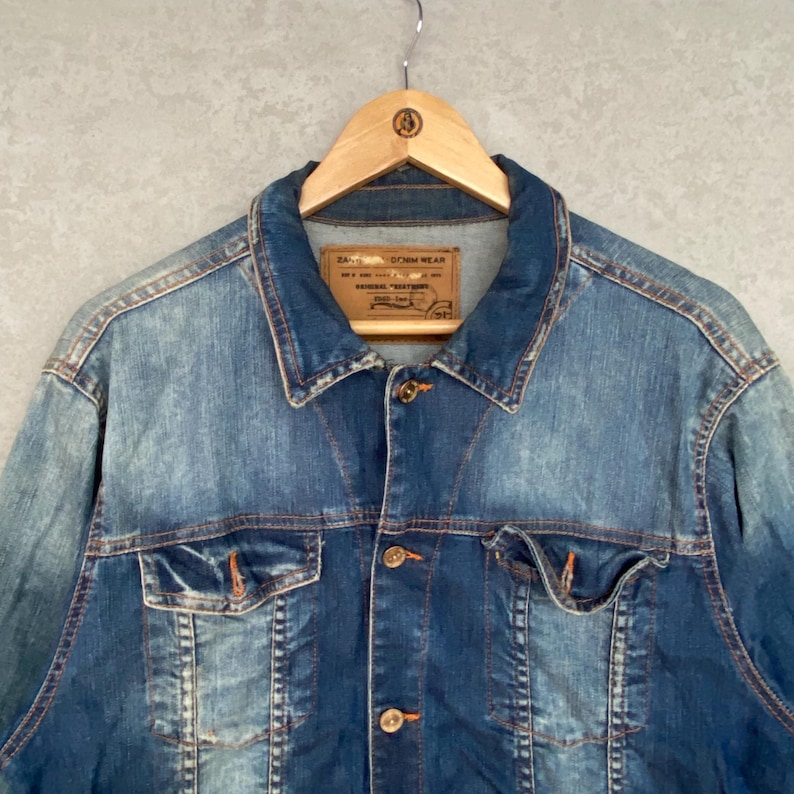 Pu&ograve; includere: Giacca di jeans blu dal design classico, con chiusura a bottoni, due tasche sul petto e colletto. La giacca &egrave; appesa a una gruccia di legno. Il denim ha un aspetto sbiadito e usurato.