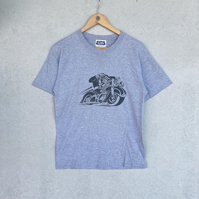 May include: A heather gray short-sleeve t-shirt featuring a black graphic print of a character riding a motorcycle with the text Von Dutch. The shirt has a crew neckline and is displayed on a wooden hanger against a neutral textured wall.