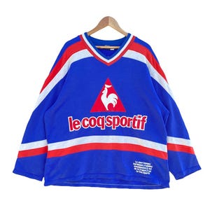 May include: Blue long-sleeve shirt with a V-neck and the text "le coq sportif". The shirt has white and red stripes across the chest and sleeves, and a rooster logo. The shirt is hanging on a wooden hanger.