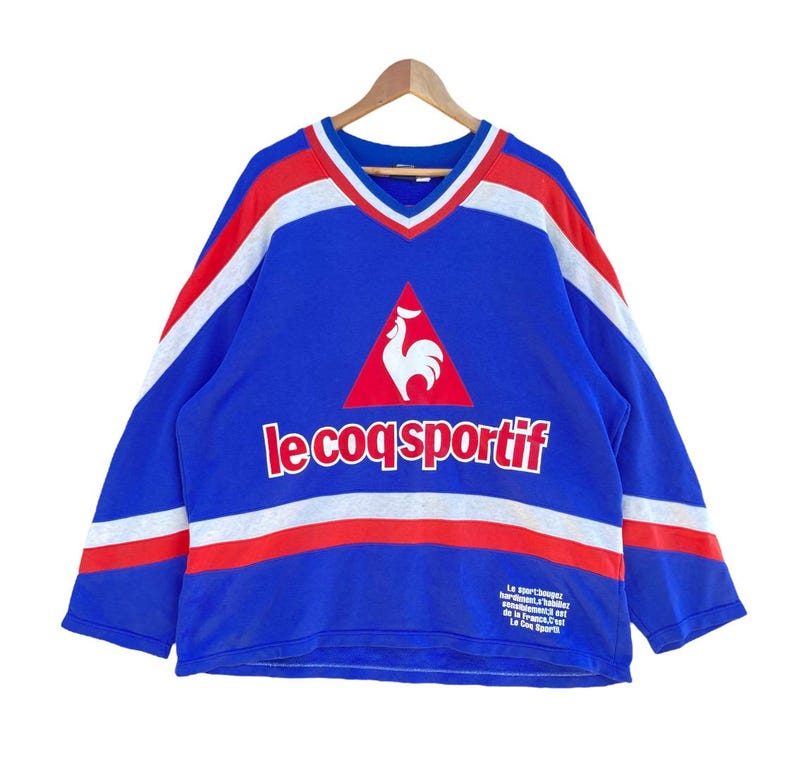 May include: Blue long-sleeve shirt with a V-neck and the text "le coq sportif". The shirt has white and red stripes across the chest and sleeves, and a rooster logo. The shirt is hanging on a wooden hanger.