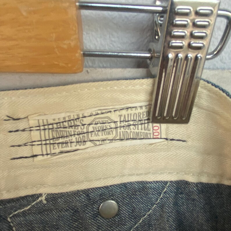 May include: Close-up of the interior waistband of denim jeans. A rectangular tag with text is stitched onto the cream-coloured fabric. The tag reads "BERY'S OVER WORKS FACTORY 1967 TAILORED FOR STYLE AND COMFORT EVERY JOB".