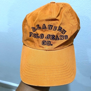 May include: An orange baseball cap with the text "R. LAUREN POLO JEANS CO." embroidered in navy blue. The cap has a curved brim and a button on top. The hat is made of a durable fabric.