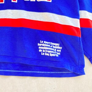 May include: Blue and white long-sleeved shirt with a red stripe across the chest. The shirt features French text, including "Le Coq Sportif."