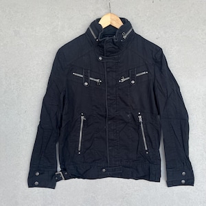 Puede incluir: Chaqueta negra con cuello alto, m&uacute;ltiples bolsillos con cremallera y detalles plateados. La chaqueta presenta una silueta entallada, una hebilla en la cintura y pu&ntilde;os abotonados. Cuelga de una percha de madera.