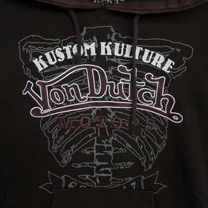 May include: A dark brown hooded sweatshirt featuring a large graphic print of a human rib cage in light grey. The text KUSTOM KULTURE is printed in white above the Von Dutch logo, with the word MOTORS in dark red below the logo.
