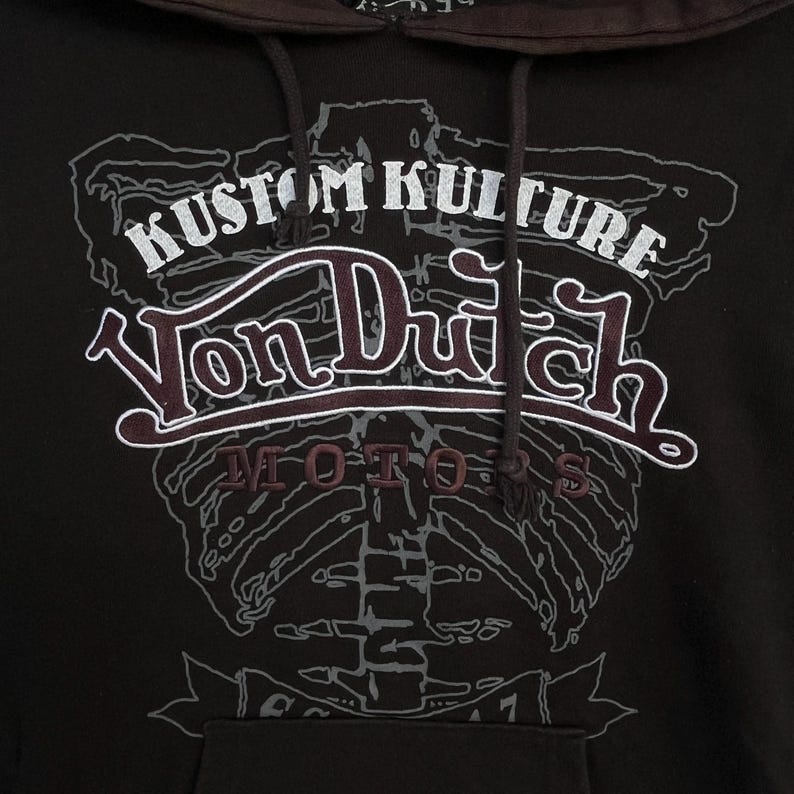 May include: A dark brown hooded sweatshirt featuring a large graphic print of a human rib cage in light grey. The text KUSTOM KULTURE is printed in white above the Von Dutch logo, with the word MOTORS in dark red below the logo.
