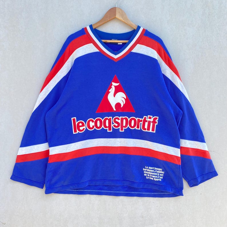 May include: Blue long-sleeve shirt with red and white stripes and a V-neck. The shirt features the Le Coq Sportif logo and text. Additional text is printed on the lower right side of the shirt.