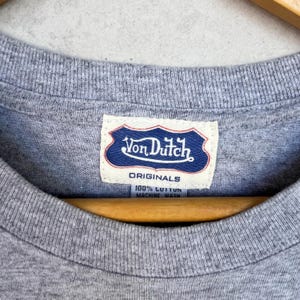May include: A heather gray t-shirt featuring a white fabric tag at the inner neckline. The tag displays a blue shield-shaped logo with the text Von Dutch, the word ORIGINALS, and the text 100% COTTON MACHINE WASH in dark blue lettering.