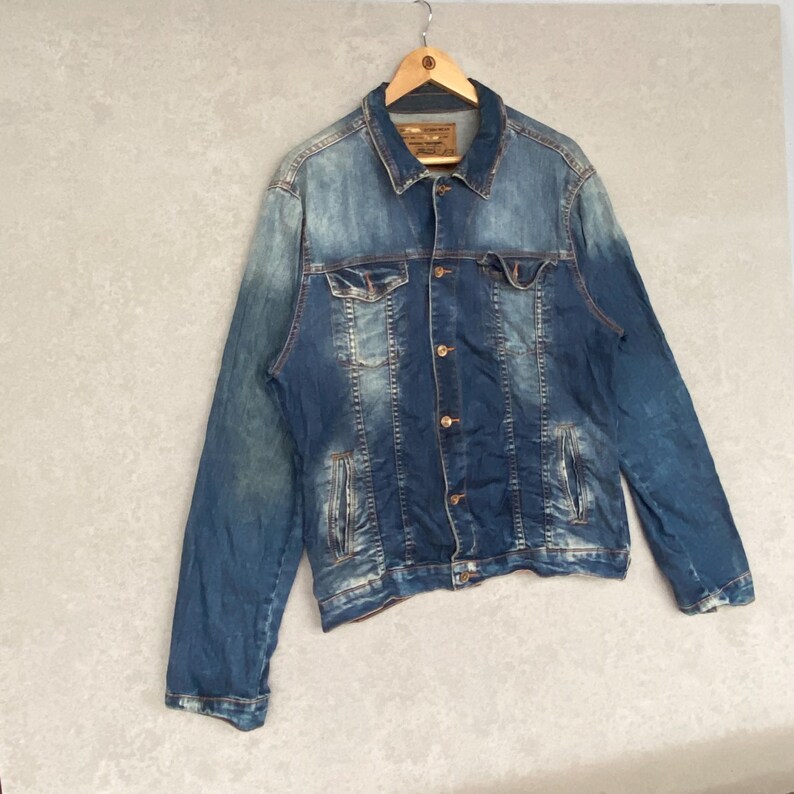 Pu&ograve; includere: Giacca di jeans blu dal design classico, con chiusura a bottoni, tasche sul petto e tasche laterali. La giacca &egrave; appesa a una gruccia di legno su uno sfondo neutro. Il denim ha un aspetto sbiadito e usurato.