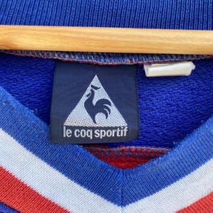 May include: Close-up of a blue Le Coq Sportif tag on a blue, red, and white striped garment. The tag features a white triangle with a black rooster and the brand name in white text. The garment is hanging on a wooden hanger.