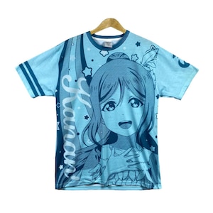 May include: Light blue t-shirt featuring a large anime-style illustration of a girl with long hair. The shirt has a crew neck and short sleeves. The word "Janan" is printed vertically on the left side of the shirt. The design includes stars and other decorative elements.