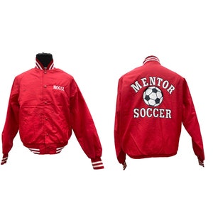 May include: Red satin varsity jacket with white embroidered lettering on the front and back. The front says "Nogsl" and the back says "Mentor Soccer" with a soccer ball graphic.