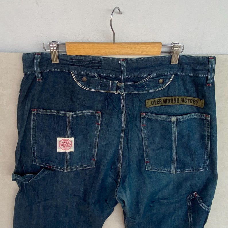 May include: Dark blue denim work trousers with red stitching. Features include multiple pockets, a belt loop, and a patch that reads "OVER WORKS FACTORY". The trousers are hanging on a wooden hanger.