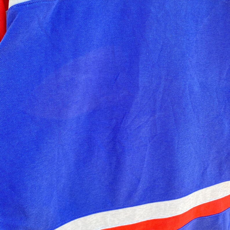 May include: Close-up of a blue, red, and white athletic shirt. The shirt is primarily blue with a white stripe and a red stripe at the bottom. The fabric appears to be a soft, knit material.