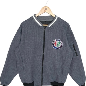 Pode incluir: Casaco bomber cinzento com fecho de correr e textura acolchoada. A gola e os punhos são listrados em branco e preto. Um patch com o logotipo da Alfa Romeo está no peito. O casaco tem um zíper preto e um pequeno bolso na manga esquerda.