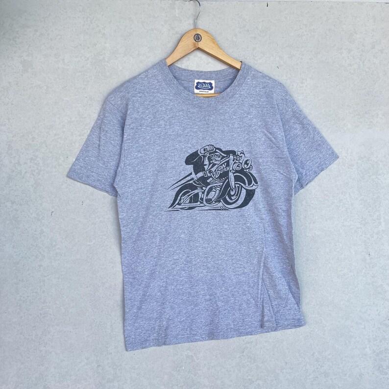 May include: A heather gray short-sleeve t-shirt featuring a black graphic print of a figure riding a motorcycle. The motorcycle has a prominent front wheel and headlight assembly. The text "VON DUTCH" is visible on the side of the motorcycle.