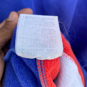 May include: Close-up of a blue, red, and white garment with a tag attached. The tag has Japanese text and care instructions. The garment appears to be made of a soft, textured material.