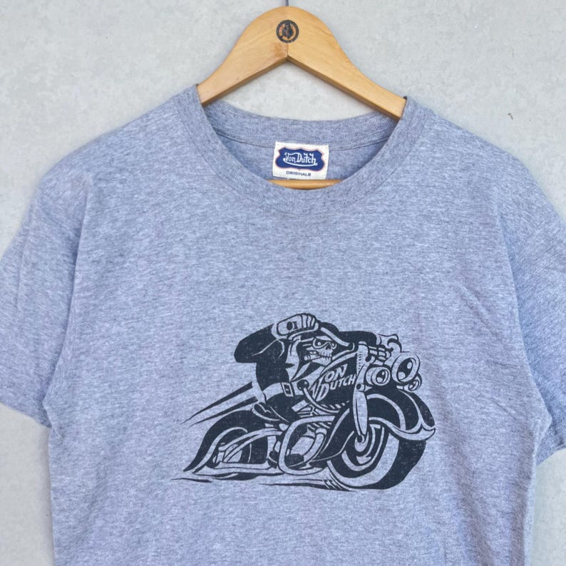 May include: A heather gray short-sleeve t-shirt featuring a black graphic print of a skeleton riding a motorcycle. The shirt has a crew neckline and a white brand label at the inner collar that reads Von Dutch Originals in blue text.
