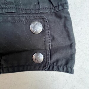 Puede incluir: Primer plano de una secci&oacute;n de tela negra con dos botones met&aacute;licos plateados. Los botones son redondos con un dise&ntilde;o en relieve detallado. La tela parece ser parte de una prenda, posiblemente una chaqueta o abrigo.