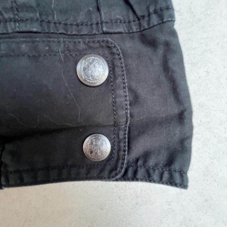 Puede incluir: Primer plano de una secci&oacute;n de tela negra con dos botones met&aacute;licos plateados. Los botones son redondos con un dise&ntilde;o en relieve detallado. La tela parece ser parte de una prenda, posiblemente una chaqueta o abrigo.