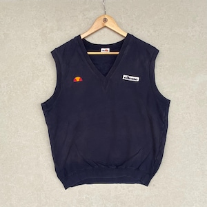 May include: A navy blue sleeveless sweater vest with a V-neck. The vest features the "ellesse" logo in white and a small orange and yellow logo on the chest. The vest is hanging on a wooden hanger.