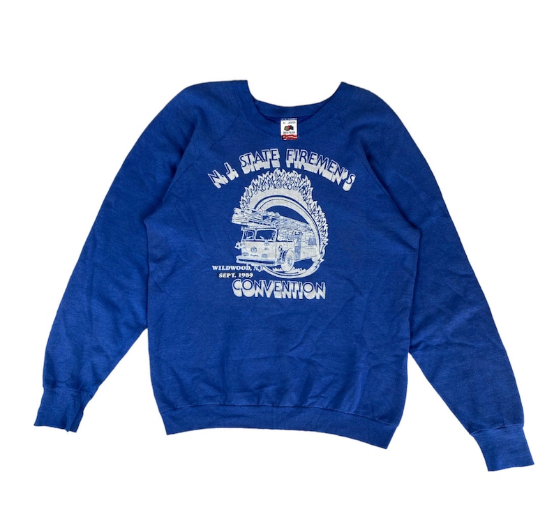 May include: Blue sweatshirt with a white graphic of a fire truck and the text "N.J. State Firemen's Convention" with the location and date "Wildwood, N.J. Sept. 1989"
